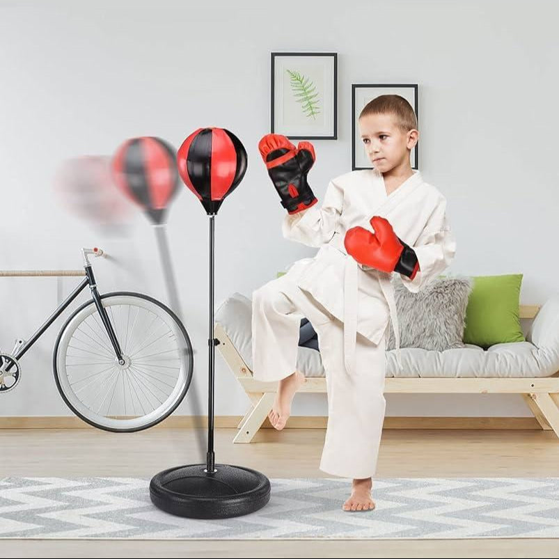 Child in a martial arts uniform with boxing gloves standing next to a punching bag in a living room.