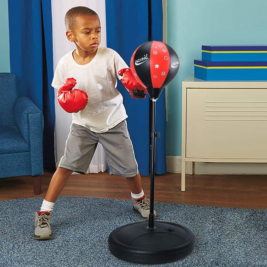 A child practicing with red boxing gloves and a black and red punching bag set, which includes a height-adjustable stand, placed on a blue carpet.